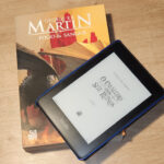 Dois livros de George R. R. Martin, "Fogo e Sangue" na versão física dando suporte à um kindle mostrando a capa de "O Cavaleiro dos Sete Reinos"