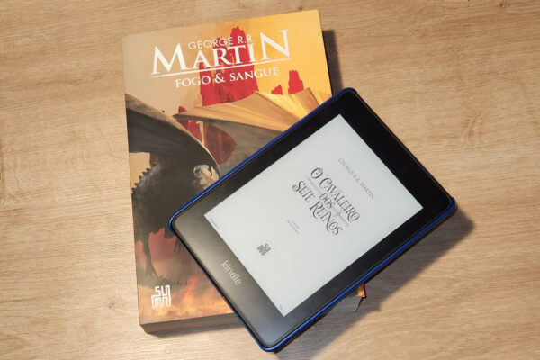 Dois livros de George R. R. Martin, "Fogo e Sangue" na versão física dando suporte à um kindle mostrando a capa de "O Cavaleiro dos Sete Reinos"