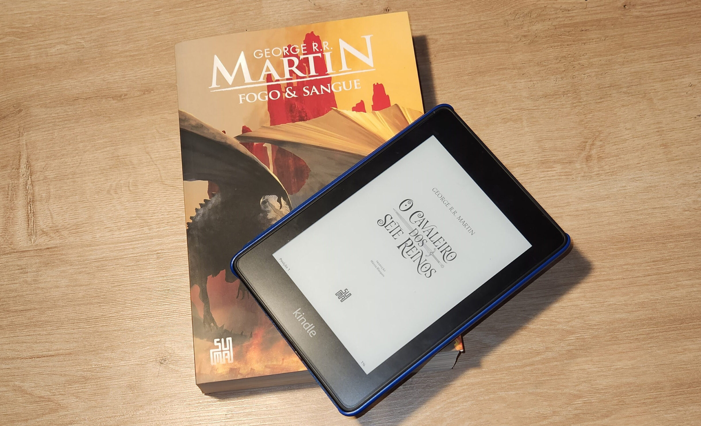 Dois livros de George R. R. Martin, "Fogo e Sangue" na versão física dando suporte à um kindle mostrando a capa de "O Cavaleiro dos Sete Reinos"